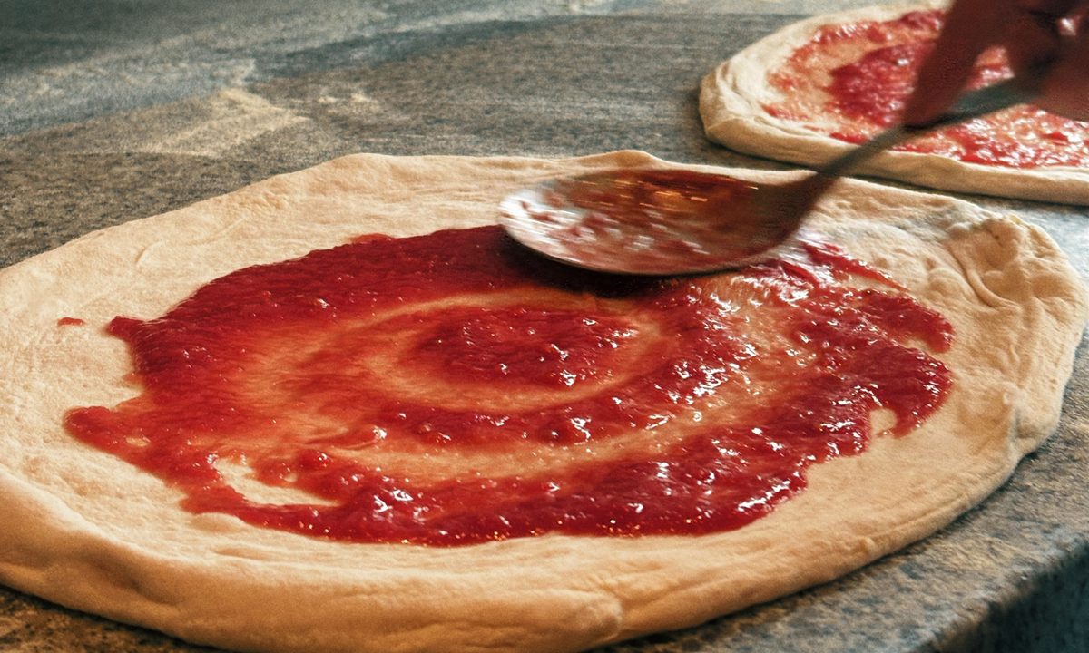 Tomato sauce being spread on pizza dough with a ladle